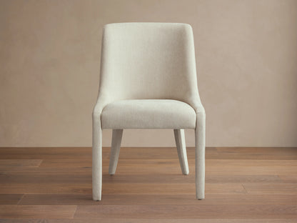 Burns Dining Chair with fully upholstered beige fabric, rounded backrest, and cushioned seat, set on a wooden floor.