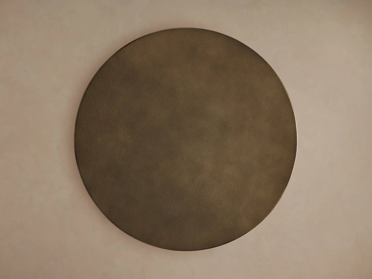 Close-up of round recycled brass-toned metal tabletop with a visible hammered texture and subtle sheen on a light background.