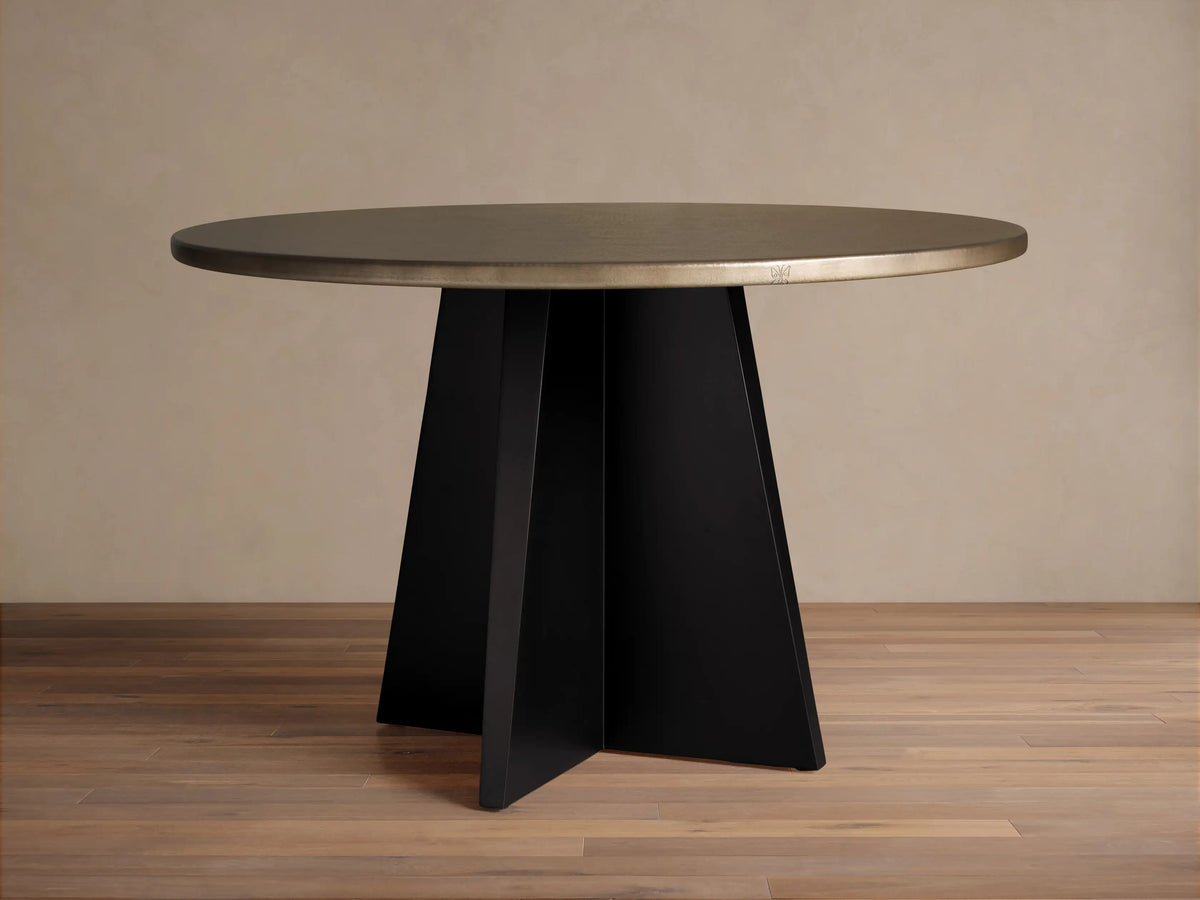 Recycled Metal Round Dining Table with a warm brassy top and a flat black vertex base, standing on a wooden floor.