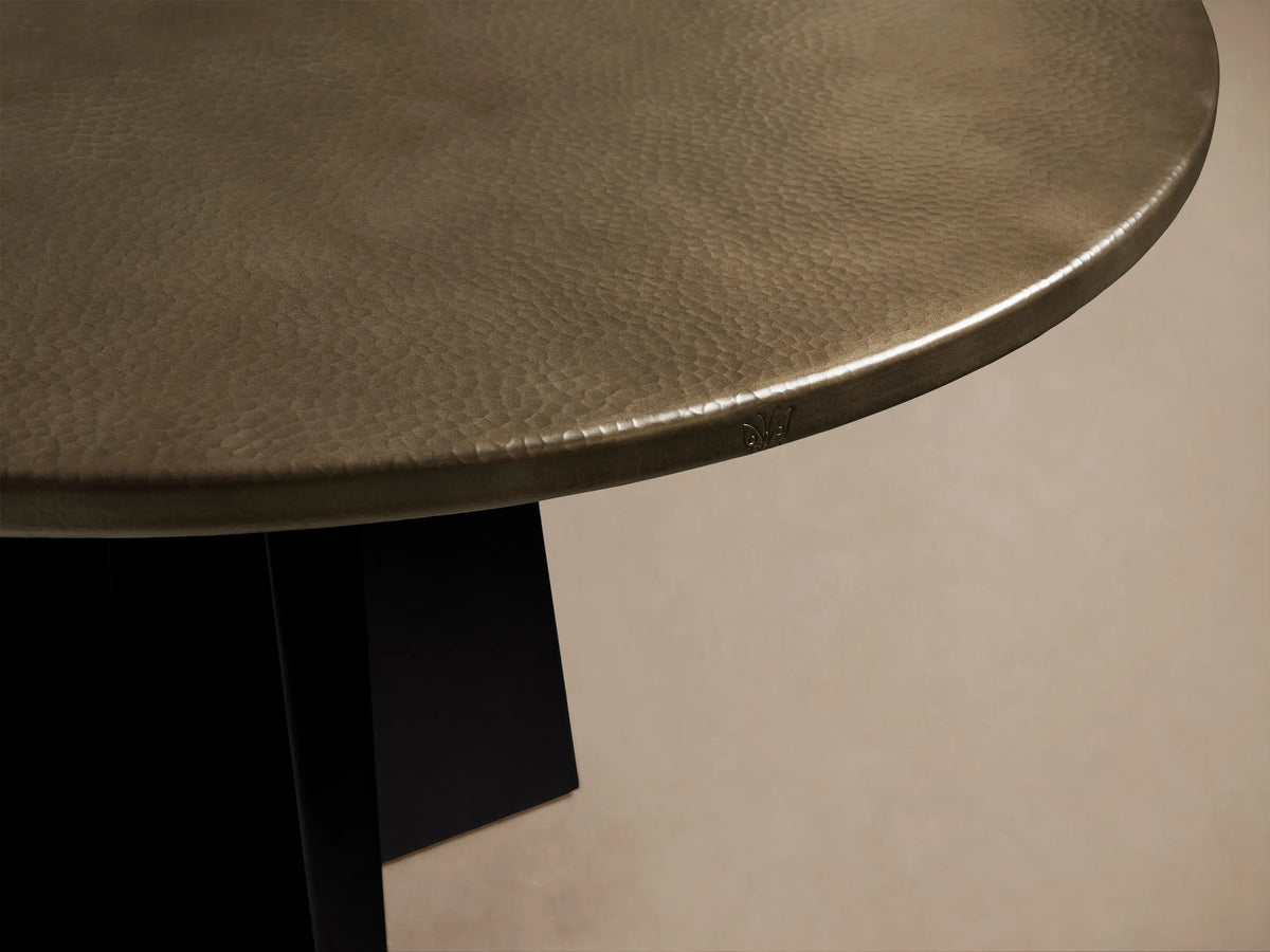 Round recycled brass-toned metal dining table with a sharp black iron Vertex Base, centered on a light wood floor.