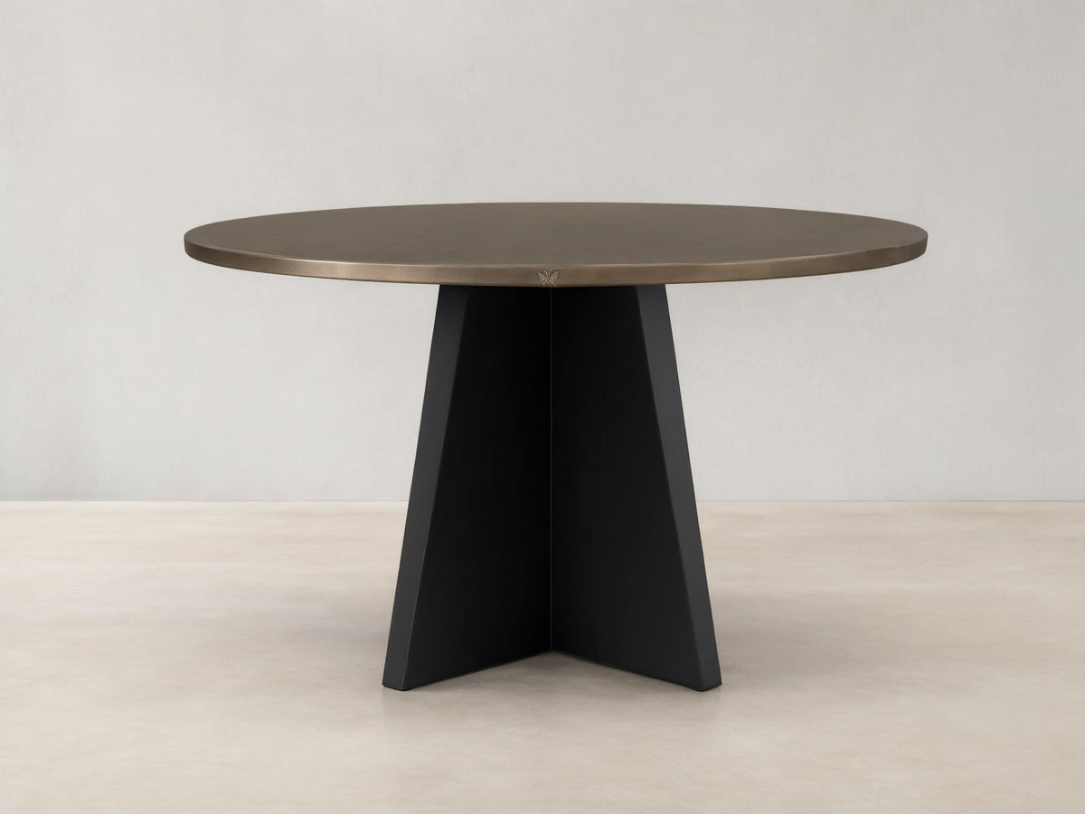 Round recycled brass-toned metal dining table with a sharp black iron Vertex Base, centered on a light wood floor.