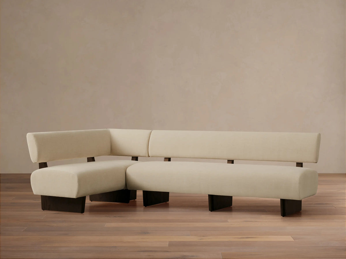 L-shaped Beau Banquette in cream fabric, featuring a sleek, minimalist design with dark wood exposed frame & legs, on wood floor.