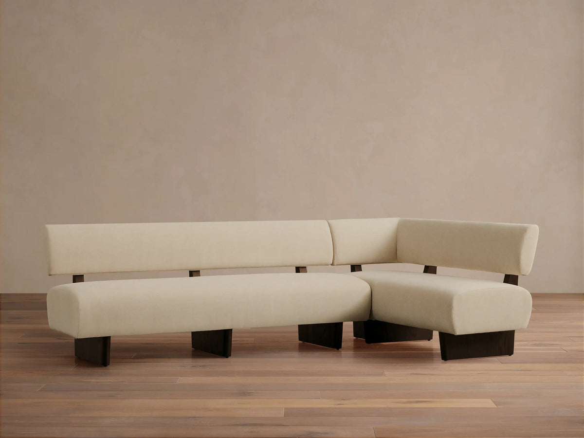 L-shaped Beau Banquette with cream upholstery and dark wood frame, sitting on a hardwood floor against a light wall.