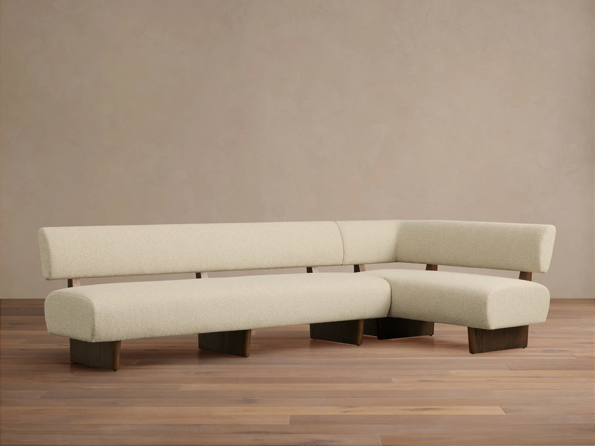 L-shaped Beau Banquette in cream textured upholstery with exposed dark wood frame and legs, on a wooden floor.