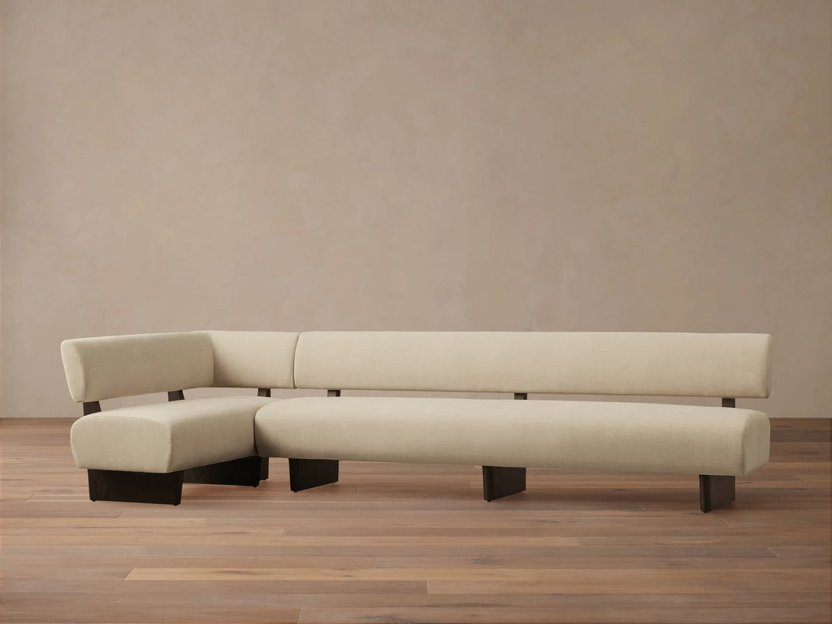 Beau Banquette, L-shaped, light cream upholstered, with exposed dark wood frame, against a light wall & wood floor.