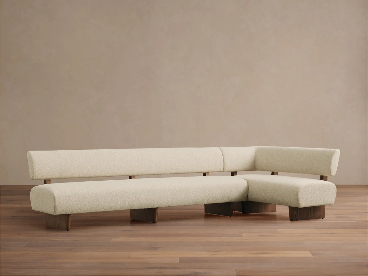 Beau Banquette, an L-shaped sofa with cream textured upholstery, dark wood base & exposed supports, on a wood floor.