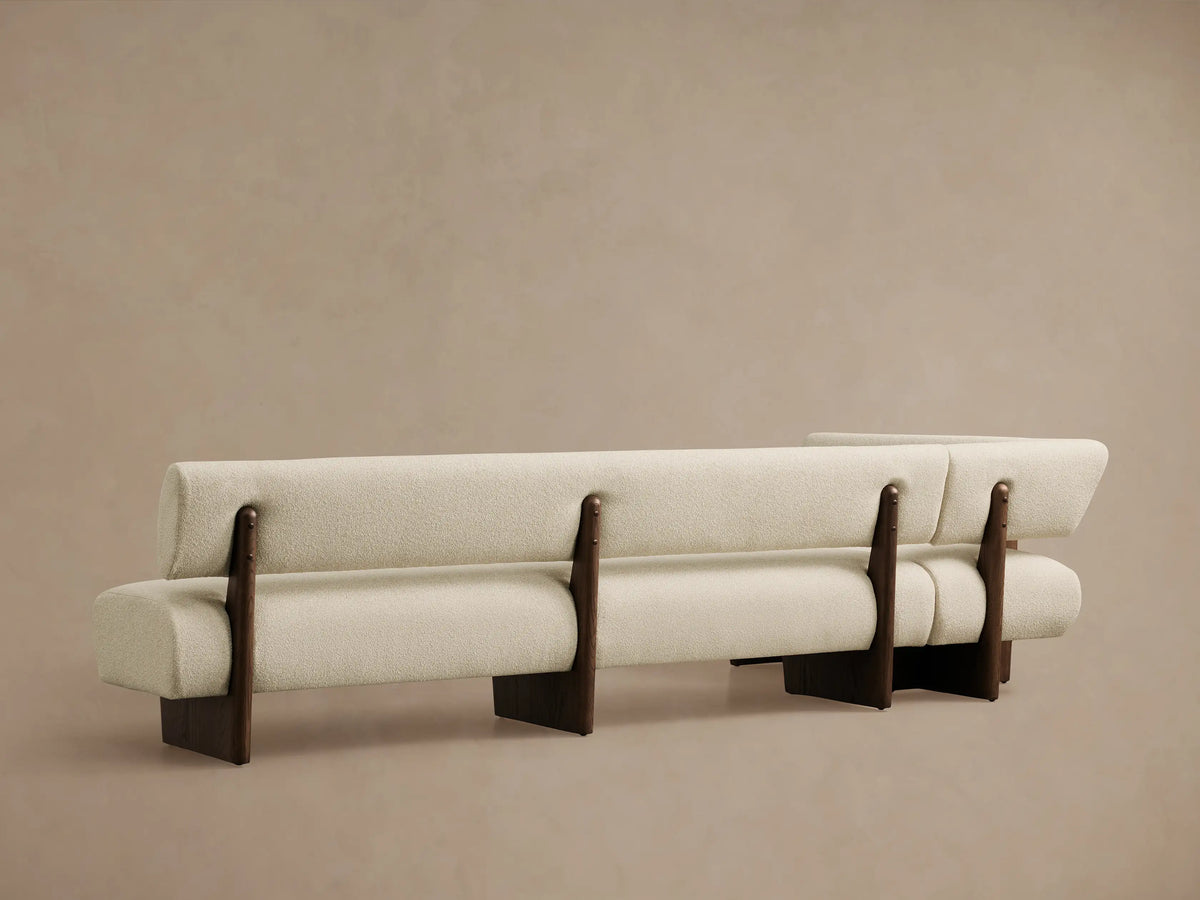 Light beige bouclv© Beau Banquette with a dark wood exposed frame, viewed from an angle showing its curved back and seat cushions.