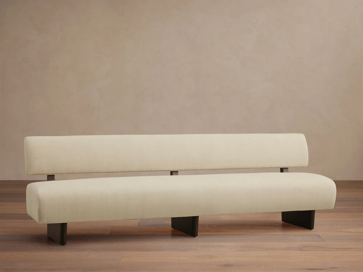 Beau Banquette, a long, cream upholstered bench with rounded seat and back cushions, supported by dark rectangular legs.