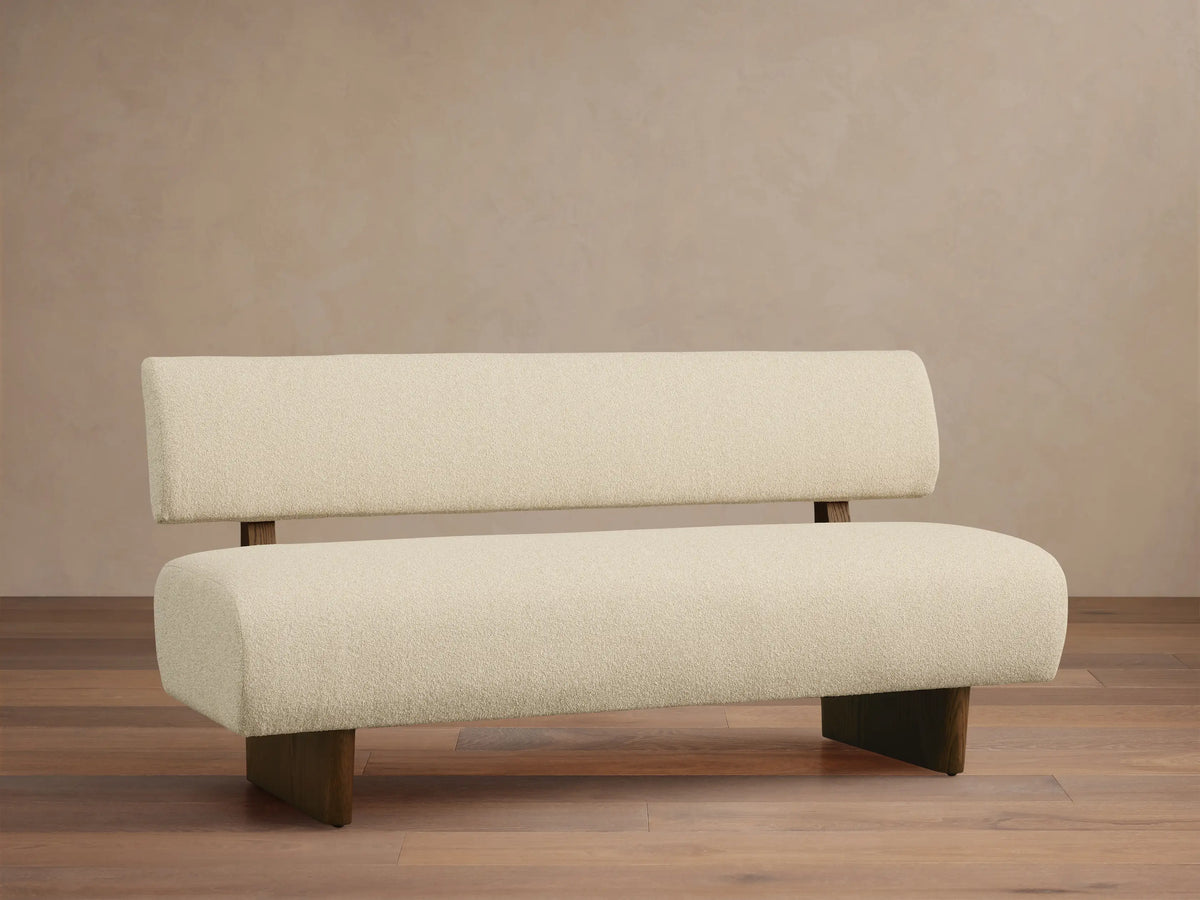 Beau Banquette: creamy white boucl?? fabric with dark wooden legs & suspended backrest, centered on a wood floor.