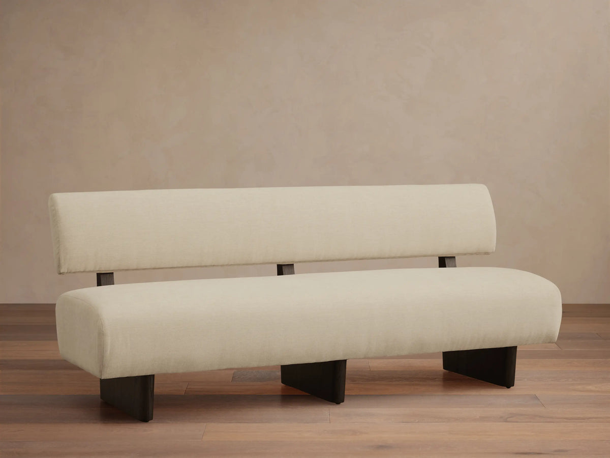 Beau Banquette: light cream upholstered with a low back and deep seat, supported by dark legs, against a beige wall & wood floor.
