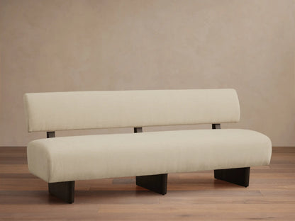 Beau Banquette: light cream upholstered with a low back and deep seat, supported by dark legs, against a beige wall & wood floor.