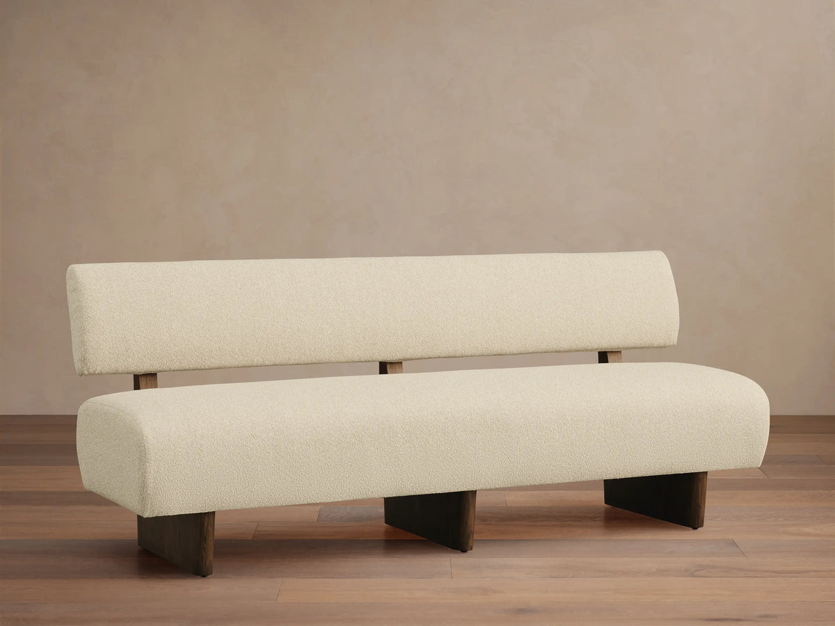 Beau Banquette, a modern bench with textured cream upholstery, dark wood legs and back supports, against a beige wall.