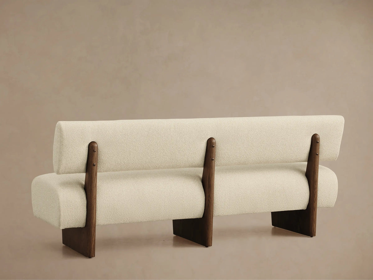 Angled top-down view of Beau Banquette. Cream textured boucle fabric with a curved backrest and dark wood frame detail.