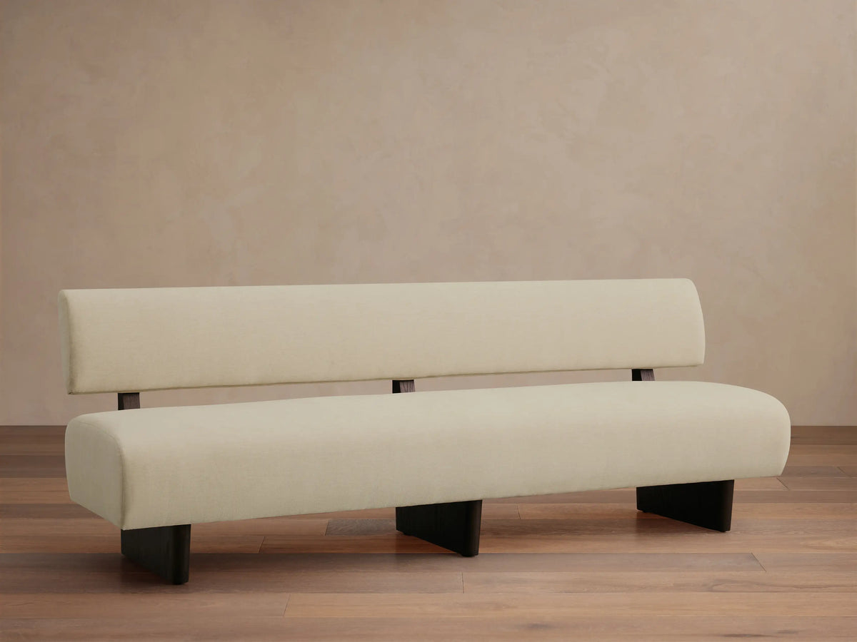 Beau Banquette: Cream upholstered modern bench with a low backrest and dark, minimalist legs, on a wood floor.