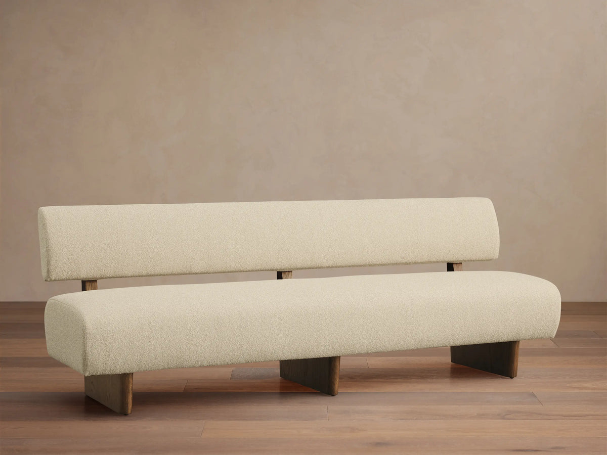 Cream upholstered Beau Banquette, modern with thick seat & backrest, dark wood supports, on a wooden floor against a wall.