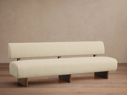 Cream upholstered Beau Banquette, modern with thick seat & backrest, dark wood supports, on a wooden floor against a wall.