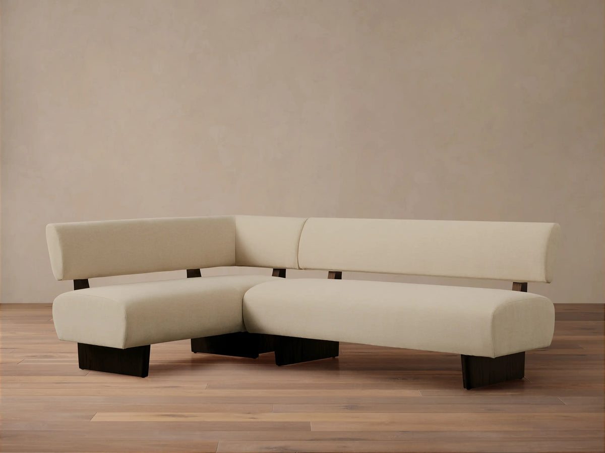 L-shaped Beau Banquette with cream upholstery and sleek dark wood base, set against a neutral wall on a wooden floor.