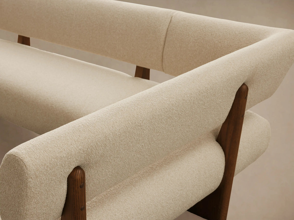 A cream, L-shaped Beau Banquette with textured upholstery and exposed dark wood frame, angled on a wooden floor.