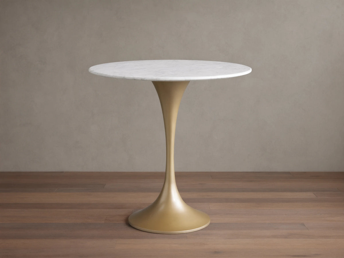 Round Saint Germain Bistro Table with a white Carrara marble tabletop and gold steel pedestal base on a wood floor background.