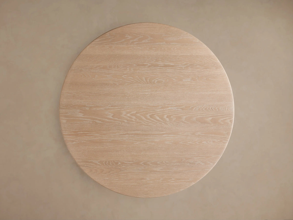 Close-up of a Caden Round Wood Dining Table. It has a light sand finish, detailed wood grain, and a thick round top on a cylindrical base.
