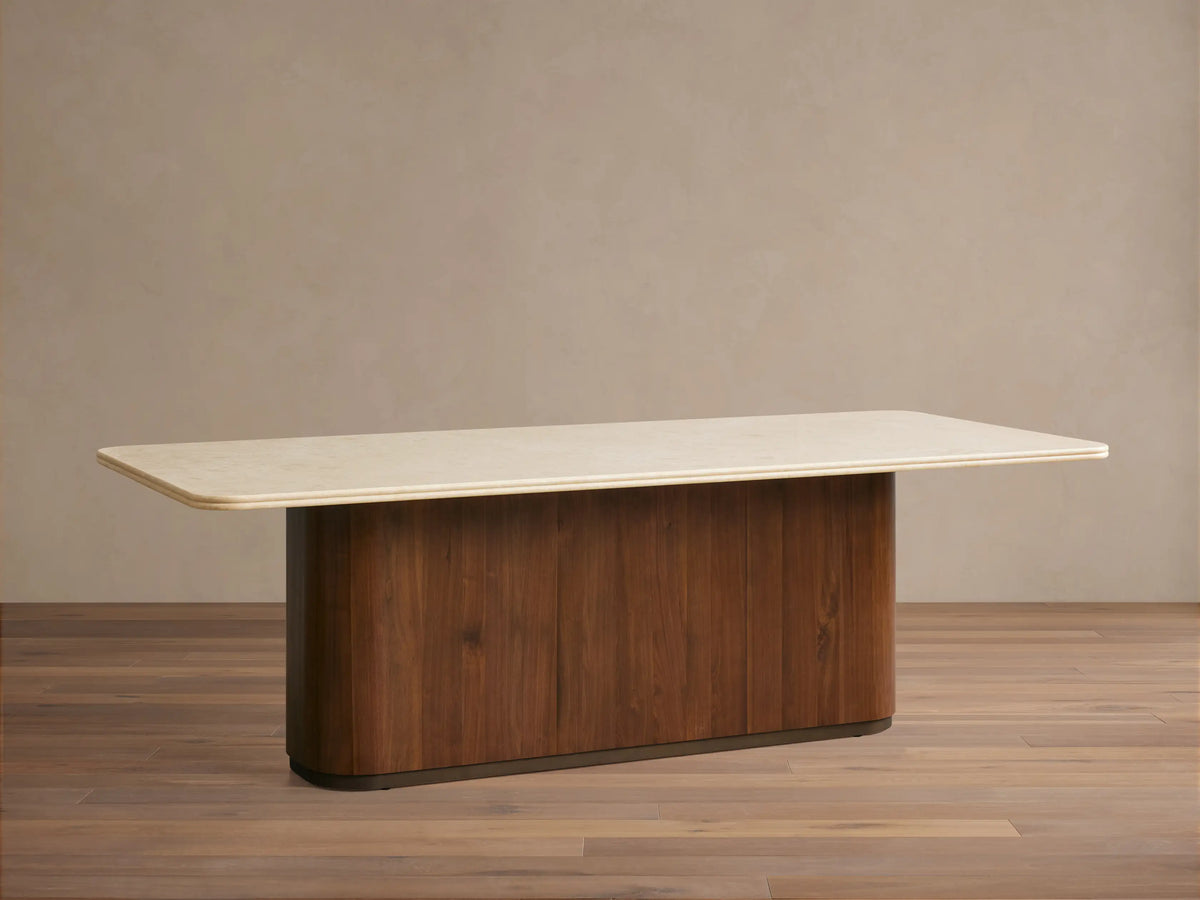 Caden Dining Table with a light, rectangular stone top and a dark walnut wood pedestal base, on a wooden floor.