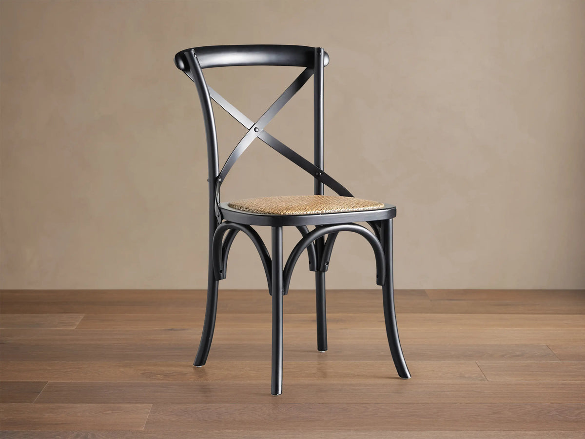 Black Cadence Dining Chair with natural rattan seat. X-back, curved legs, angled on a brown wood floor against a beige wall.