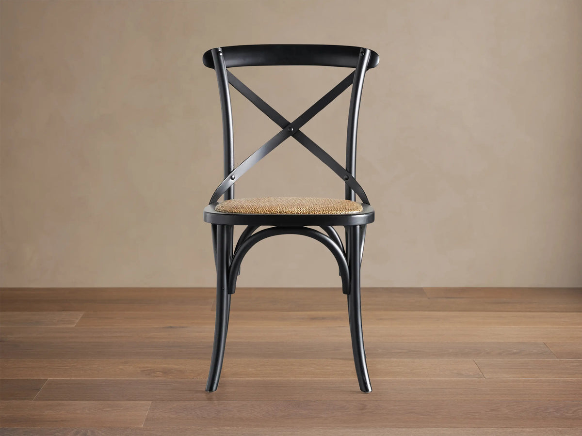 Black Cadence Dining Chair with natural rattan seat. X-back, curved legs, angled on a brown wood floor against a beige wall.