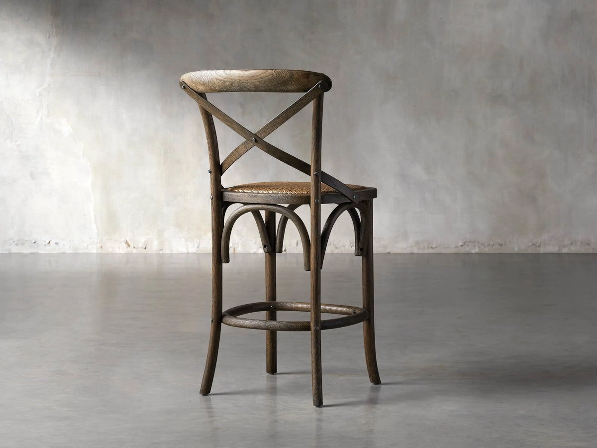 Close-up of Cadence Stool's dark brown weathered oak cross-back, showing wood grain and metal rivets against a dark background.