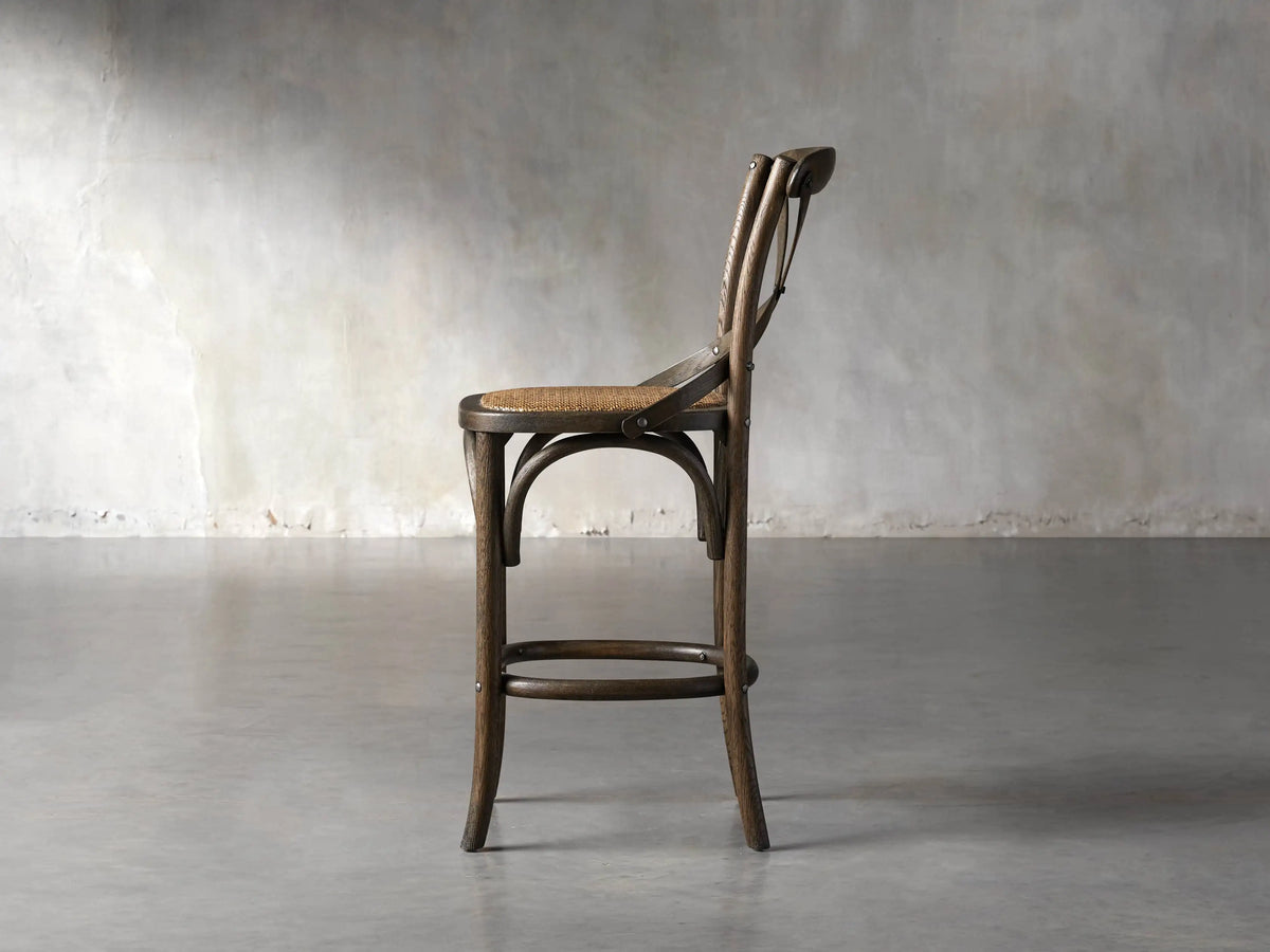 Cadence Stool, dark weathered wood, with curved X-back, round footrest, and woven natural rattan seat.