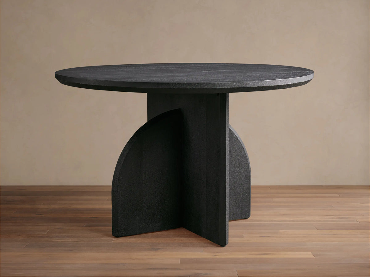 Calderone Dining Table: Round black mango wood top with sandblasted finish, unique geometric base, matte. On wood floor.