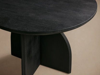 Calderone Dining Table: Round black mango wood top with sandblasted finish, unique geometric base, matte. On wood floor.