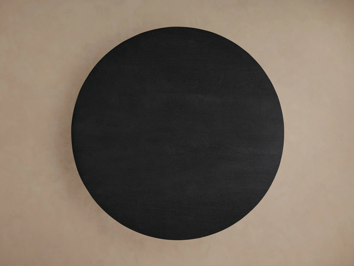 Calderone Dining Table: Angled close-up of a round sandblasted black mango wood top, thick curved & angular base.
