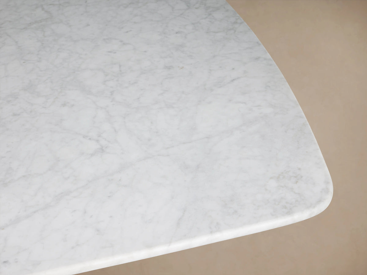Saint Germain Stone Dining Table with a long, white Carrara marble top and a rectangular, bronze-colored Clement base on a wood floor.