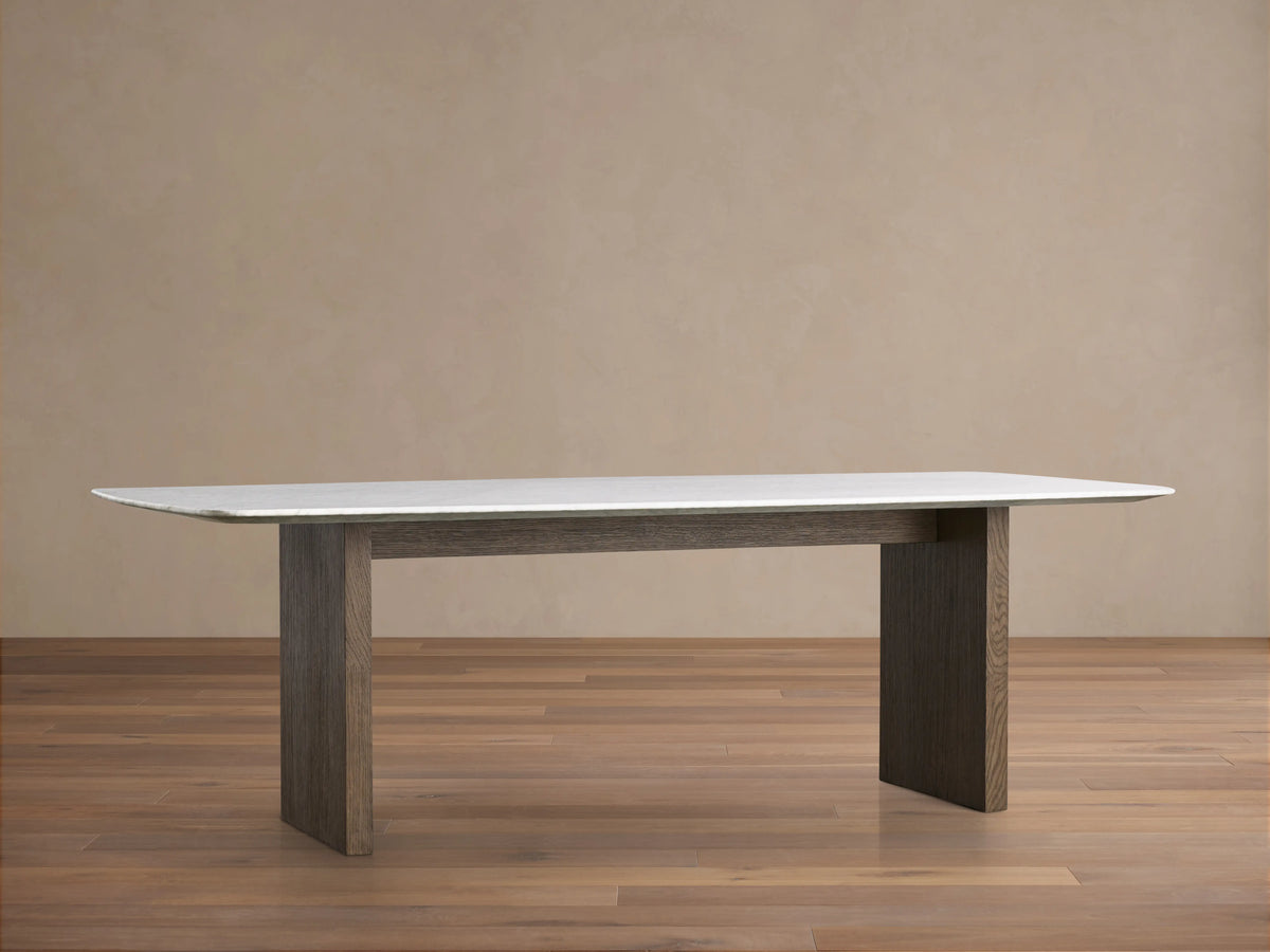 Saint Germain dining table with a Carrara marble top and two thick wood legs in a chestnut finish on a wooden floor.