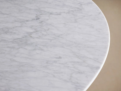 Round stone dining table with a smooth white Carrara marble top and a sturdy, flat, golden Clement base. Set on a warm wooden floor against a neutral beige wall.