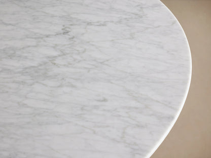 Round dining table with a Carrara marble top and a wood base in a natural finish, set against a wooden floor and neutral wall.