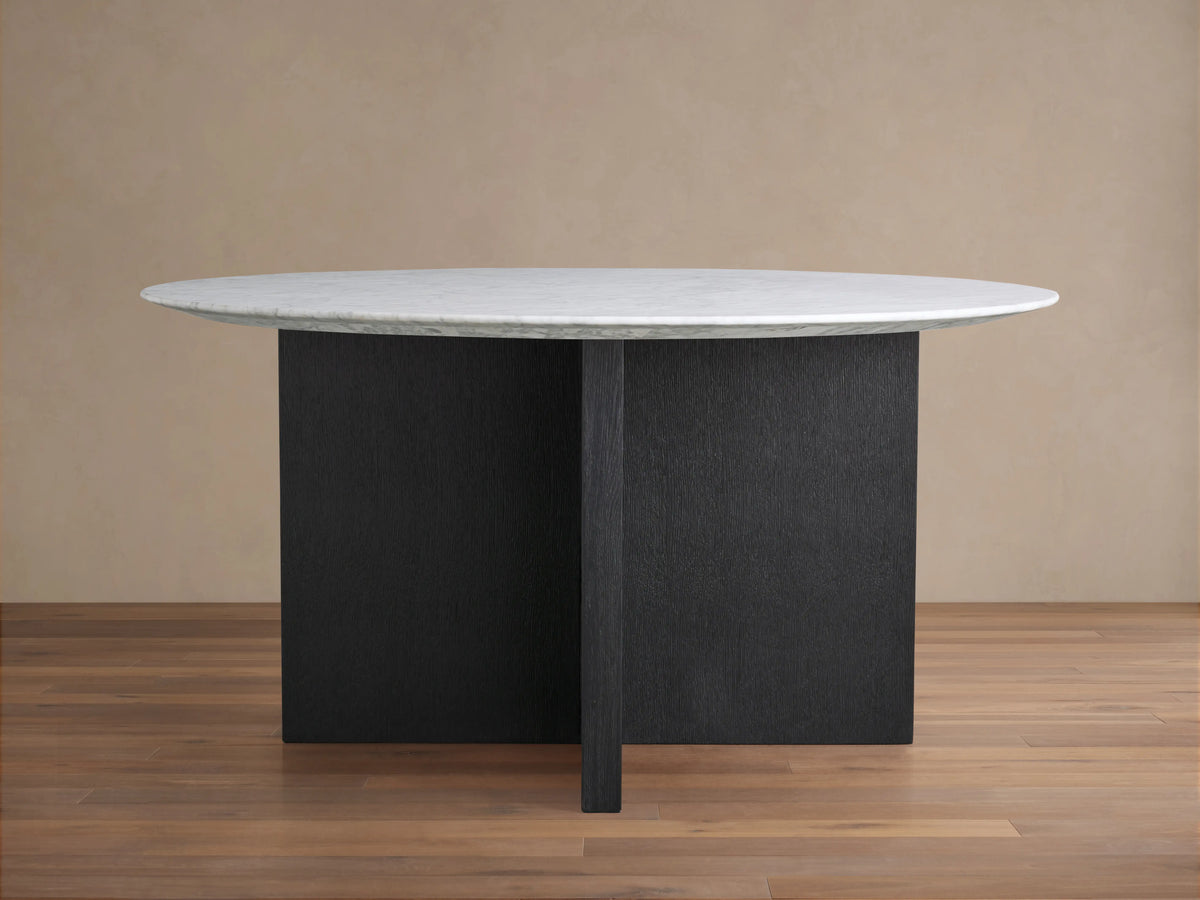 Round dining table with a smooth, white stone top and a sturdy, cross-shaped dark wood base. Positioned on a wooden floor against a neutral wall.