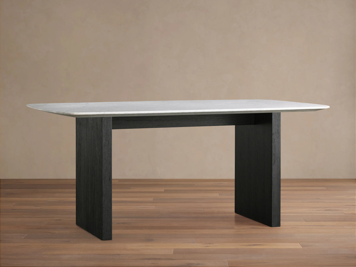 Saint Germain Stone Top Dining Table with a light marble top and dark wooden base, set on a wooden floor, against a beige wall.