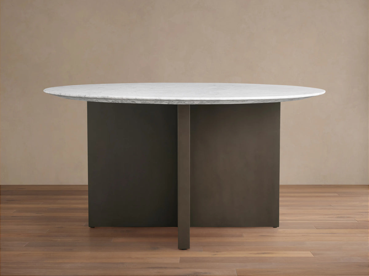 Round dining table with a white Carrara marble top and a V-shaped dark brown base on a wooden floor against a beige wall.