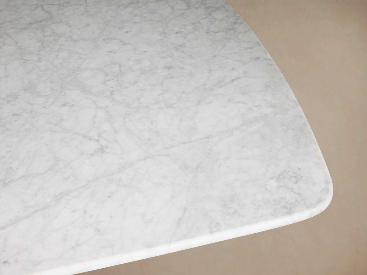 The image shows a close-up of the Saint Germain Stone Dining Table with a Carrara marble top. The surface features subtle gray veining on a light background and smooth, curved edges.