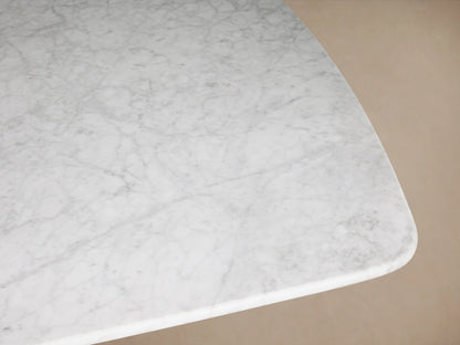 The image shows a close-up of the Saint Germain Stone Dining Table with a Carrara marble top. The surface features subtle gray veining on a light background and smooth, curved edges.