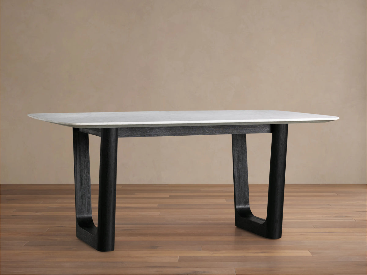A rectangular dining table with a white Carrara marble top and a thick black wooden base, set on a wooden floor with a beige wall background.