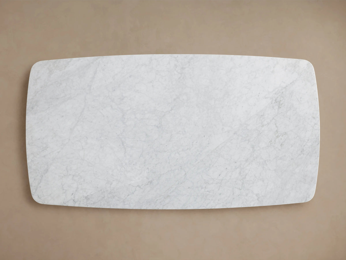 Saint Germain Stone Dining Table with a rectangular Carrara marble top, featuring a unique pattern. It has a dark wood Rennes base in an arch shape, on a wooden floor.