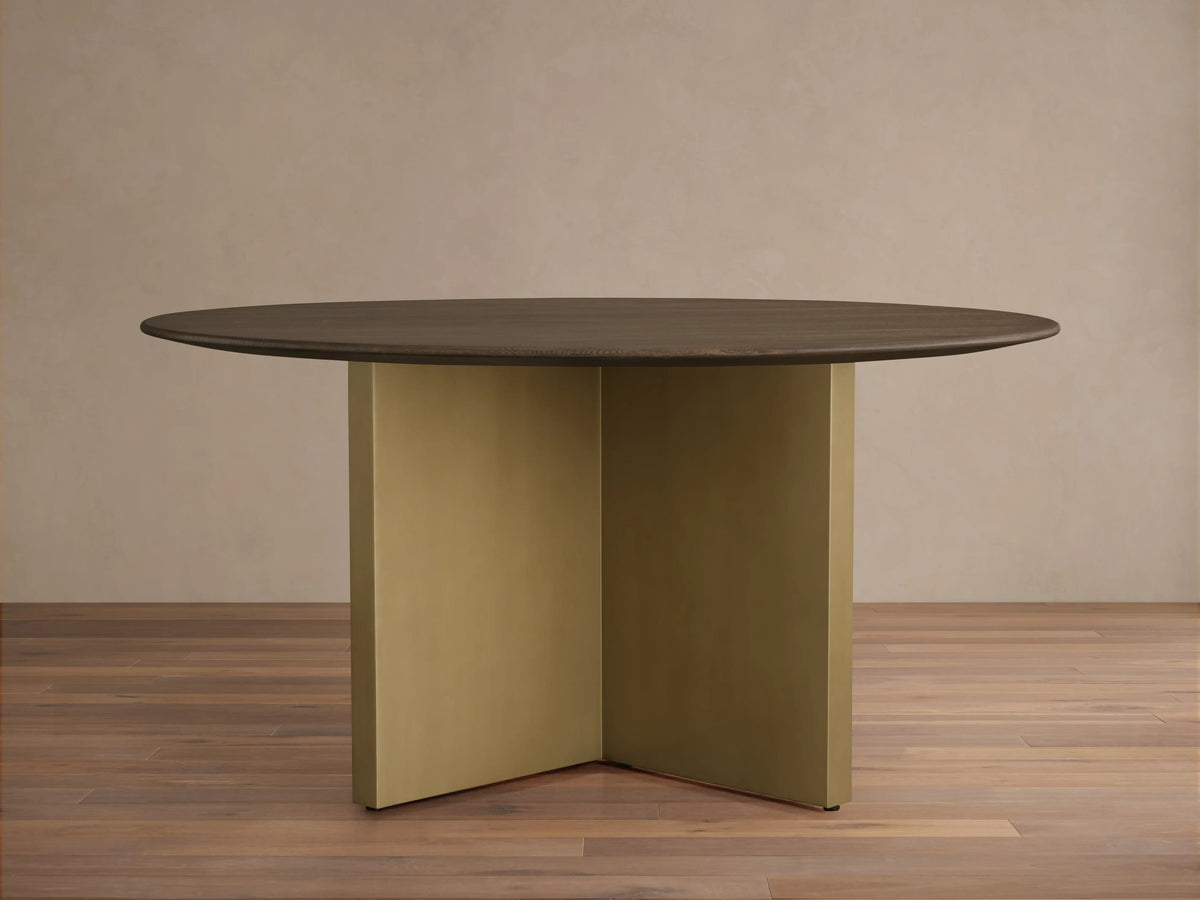 Round dining table with a solid oak chestnut finish top and a V-shaped antique brass steel base on a wooden floor.