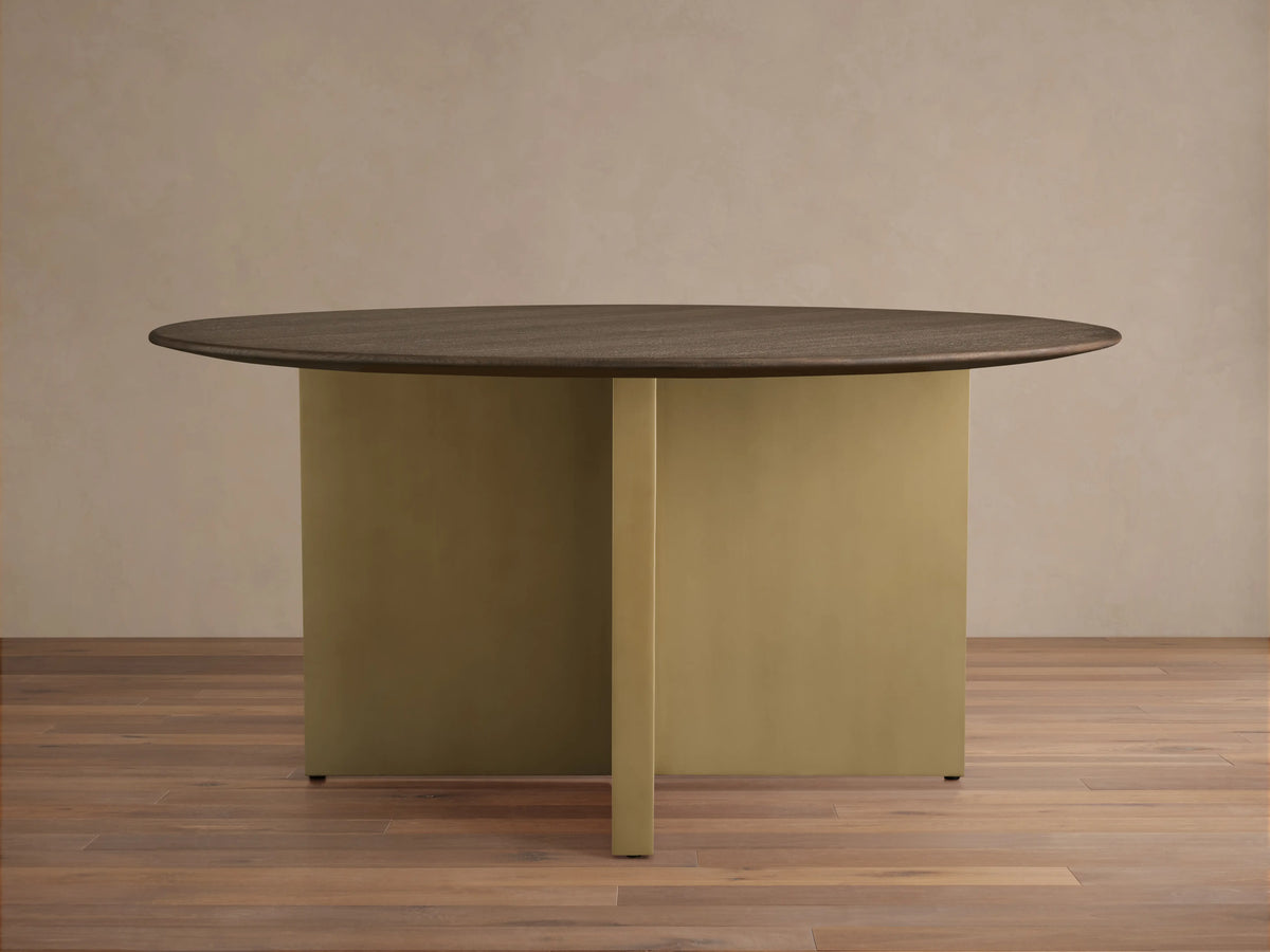 Round dining table with a solid oak chestnut finish top and a V-shaped antique brass steel base on a wooden floor.