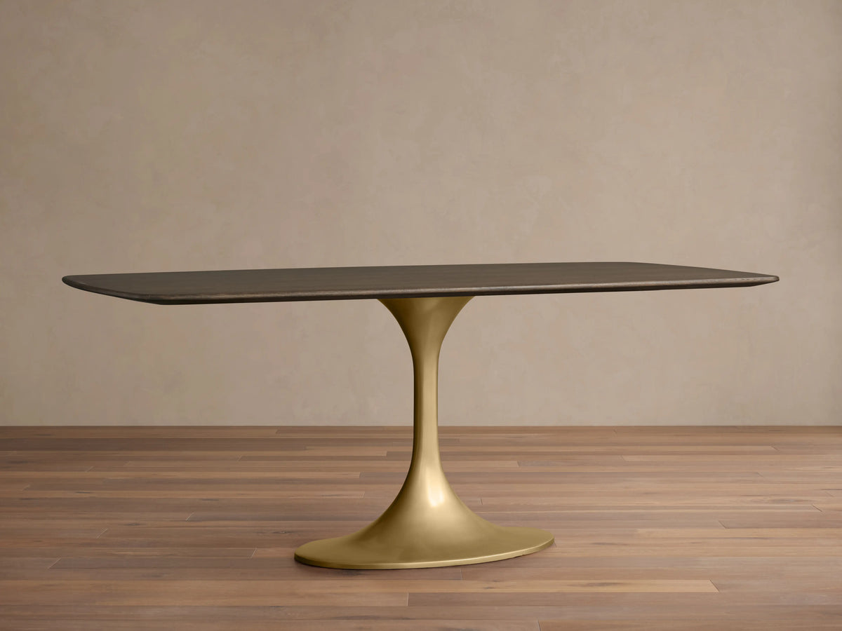 Rectangular Saint Germain wood dining table with dark oak top and elegantly curved gold Launette steel base on wood floor.
