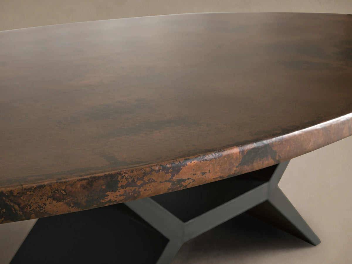 Recycled Metal Dining Table with a textured, oval copper-toned top and a sculptural black Vertex base on a wooden floor.