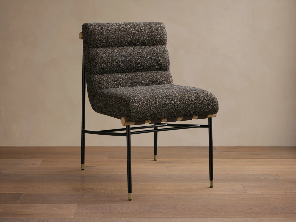 Front view of an armless accent chair featuring dark speckled boucle fabric, channel-tufted back, black metal legs with gold caps, and light wood accents.