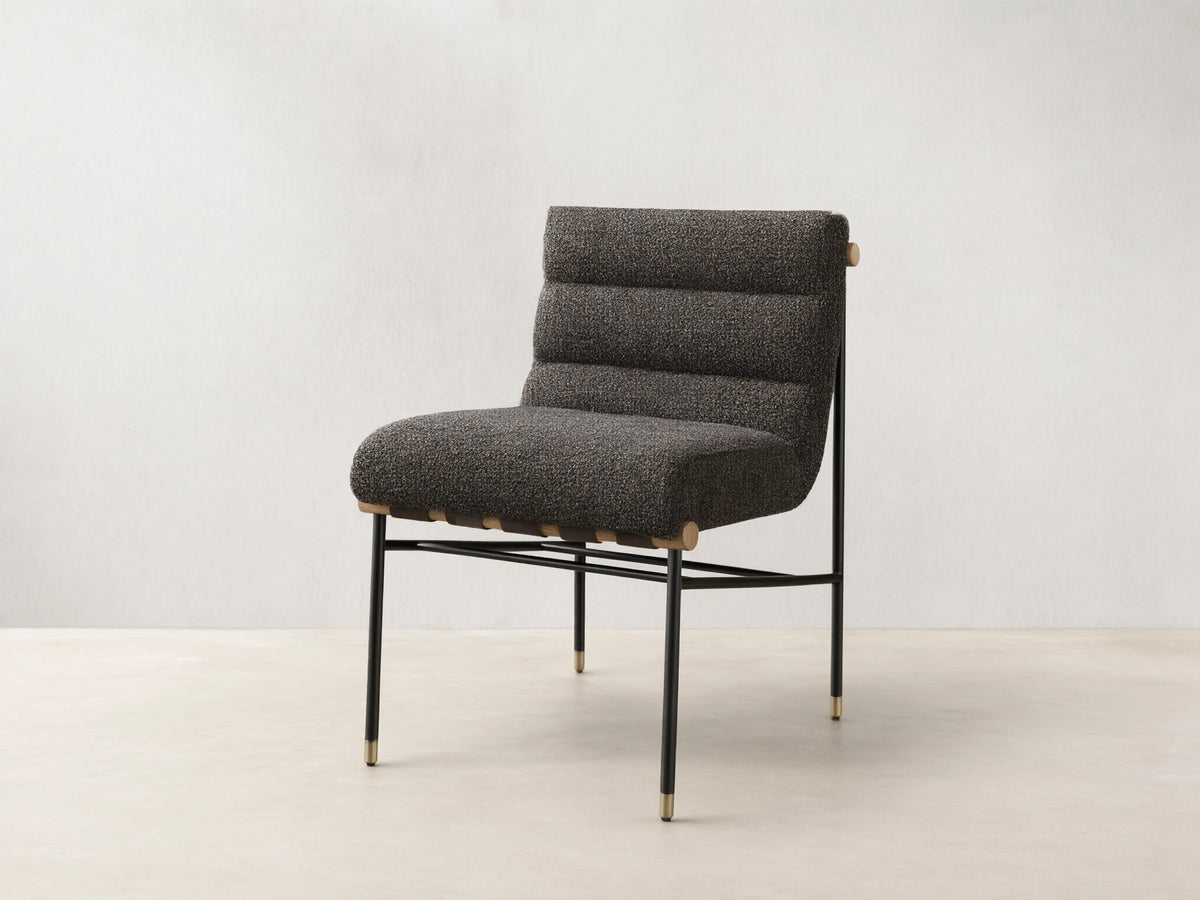 Front view of an armless accent chair featuring dark speckled boucle fabric, channel-tufted back, black metal legs with gold caps, and light wood accents.