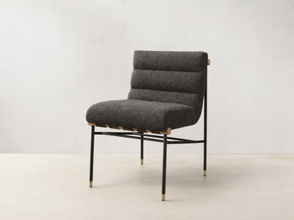 Front view of an armless accent chair featuring dark speckled boucle fabric, channel-tufted back, black metal legs with gold caps, and light wood accents.